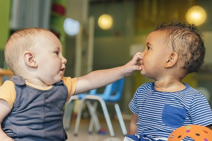 Two infants sitting together with one child reaching across to touch the nose of the other child