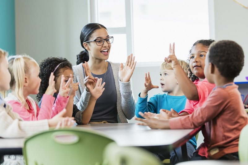 Educator sitting at a table with children playing a counting game with hands up in the air