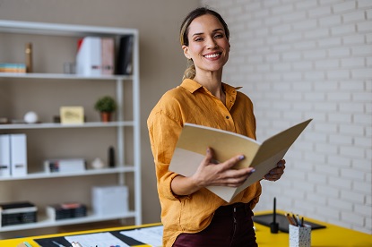 A happy and relaxed educator holding open a notebook while leaning on the back of the desk