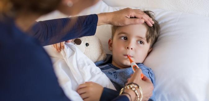 A child in bed with their soft toy and getting their temperature checked orally and with an adult's hand checking the child's forehead