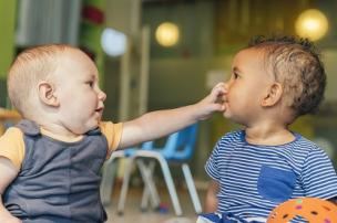 Two infants sitting together with one child reaching across to touch the nose of the other child