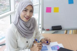 Educator sitting at a desk with paperwork and post it notes on a whiteboard in the background